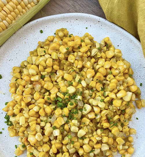 A plate of buttery corn kernels topped with chopped chives, beside a fresh cob of corn
