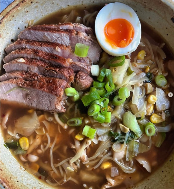 A bowl of ramen with sliced roast meat, a jammy soft-boiled egg, scallion greens, corn and enoki mushrooms