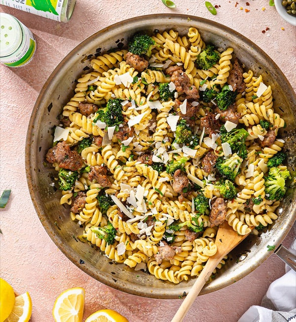 A skillet of fusilli with broccoli, crumbled sausage and shaved parmesan, beside a jar of Gourmend powder and lemon halves