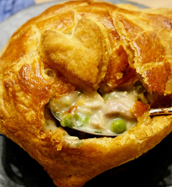A single-serve chicken pot pie with a puff pastry lid and a heart cut-out, opened to a creamy filling with peas