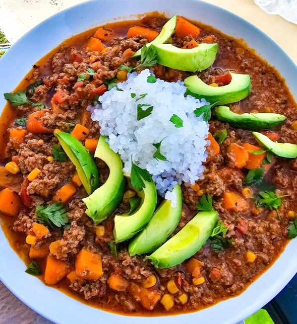 A bowl of lamb chili with sweet potato, corn and tomato, topped with sliced avocado, cilantro and rice