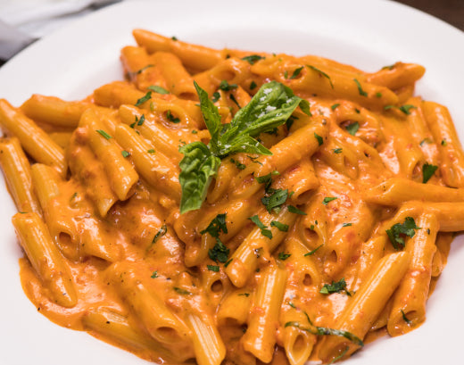 A white bowl of penne in a creamy tomato sauce, scattered with chopped parsley and topped with fresh basil