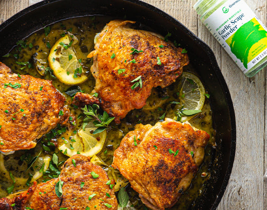 Golden-seared chicken thighs in a cast-iron skillet with lemon slices, capers and fresh herbs, beside a jar of Gourmend Garlic Scape Powder