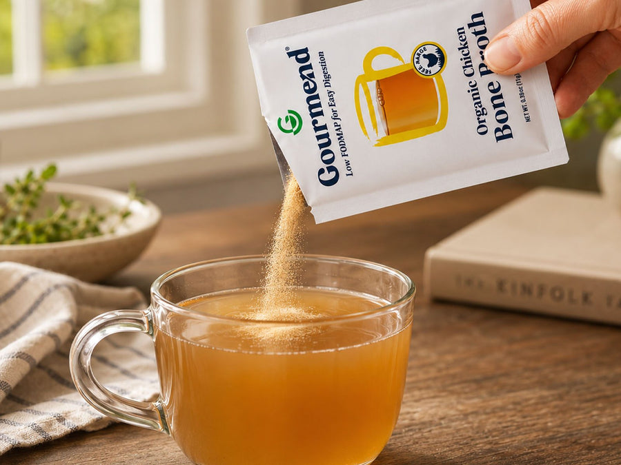 A hand pouring a packet of Gourmend chicken bone broth powder into a glass mug of hot broth on a wooden table