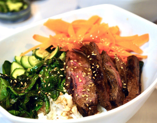 Sliced medium-rare steak with sesame seeds over rice, with sautéed spinach, cucumber and shredded carrot