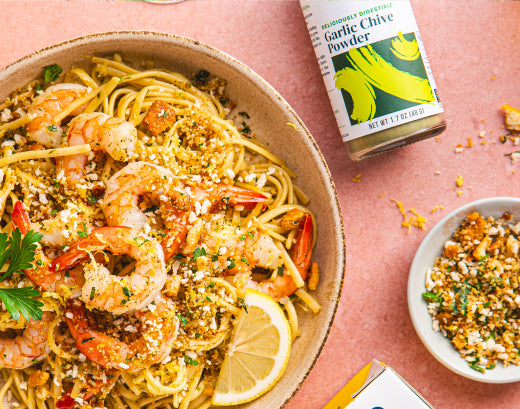A bowl of shrimp scampi linguine with herbs and toasted crumbs, beside a jar of Gourmend Garlic Chive Powder