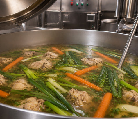 Broth simmering in a large steam kettle with chicken, carrots, leek greens and scallion tops